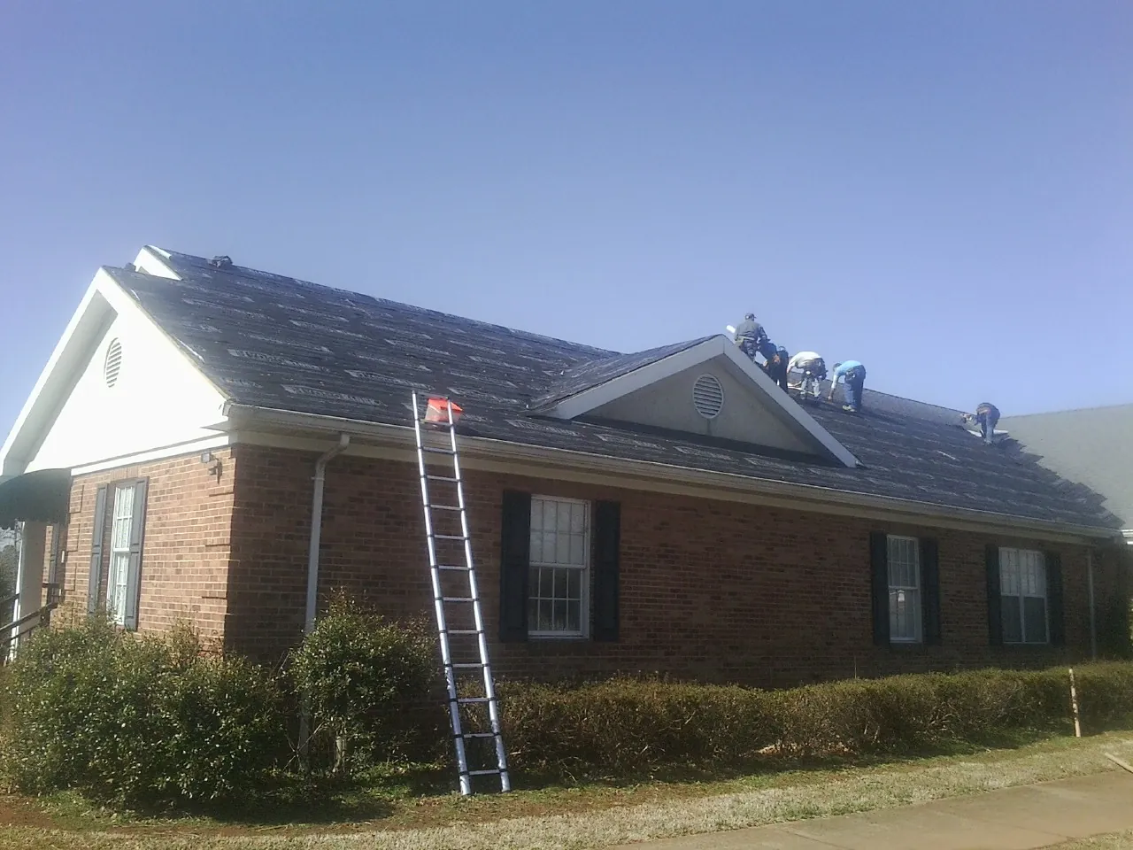 Crew tearing shingles off a brick ranch roof with a ladder set — Emergency Roof Repair work by our crew in Stone Ridge