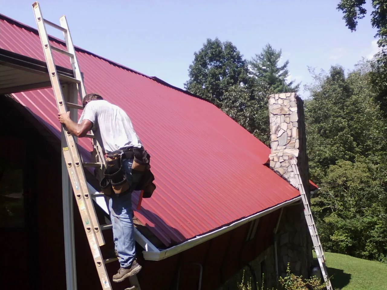 Roofer on a ladder beside a red metal roof and stone chimney — Soffit & Fascia Repair by Roofing Brothers in Stone Ridge